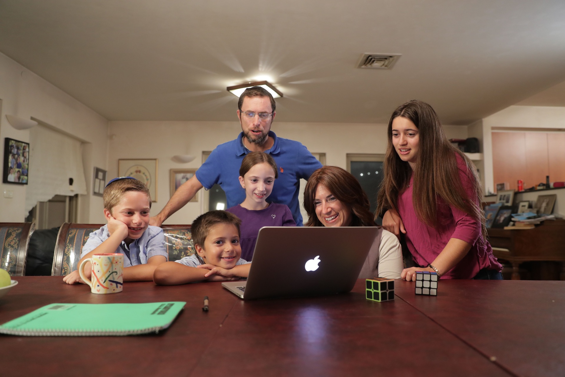 A whole family of six gathered around a MacBook at the dining table