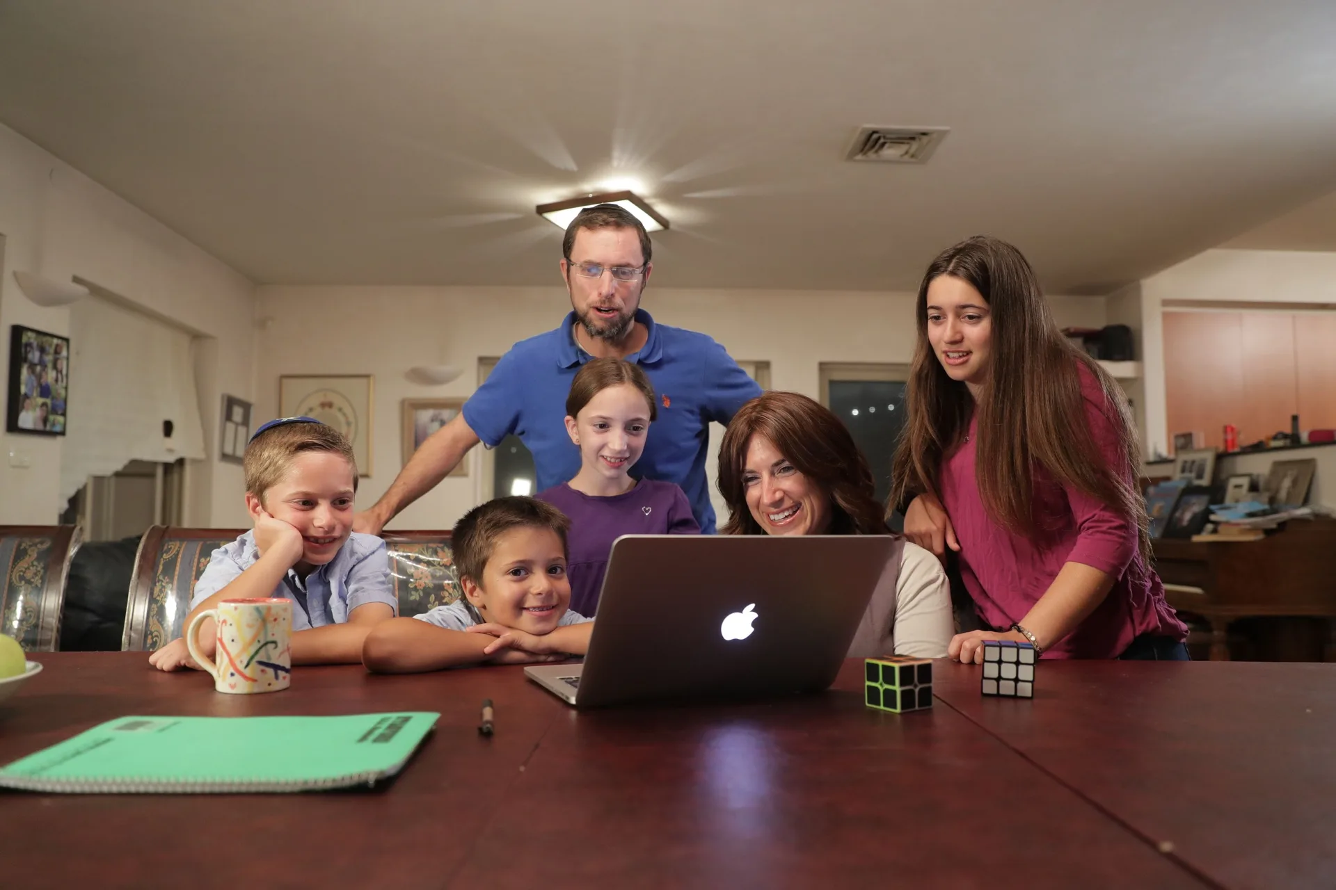A whole family of six gathered around a MacBook at the dining table